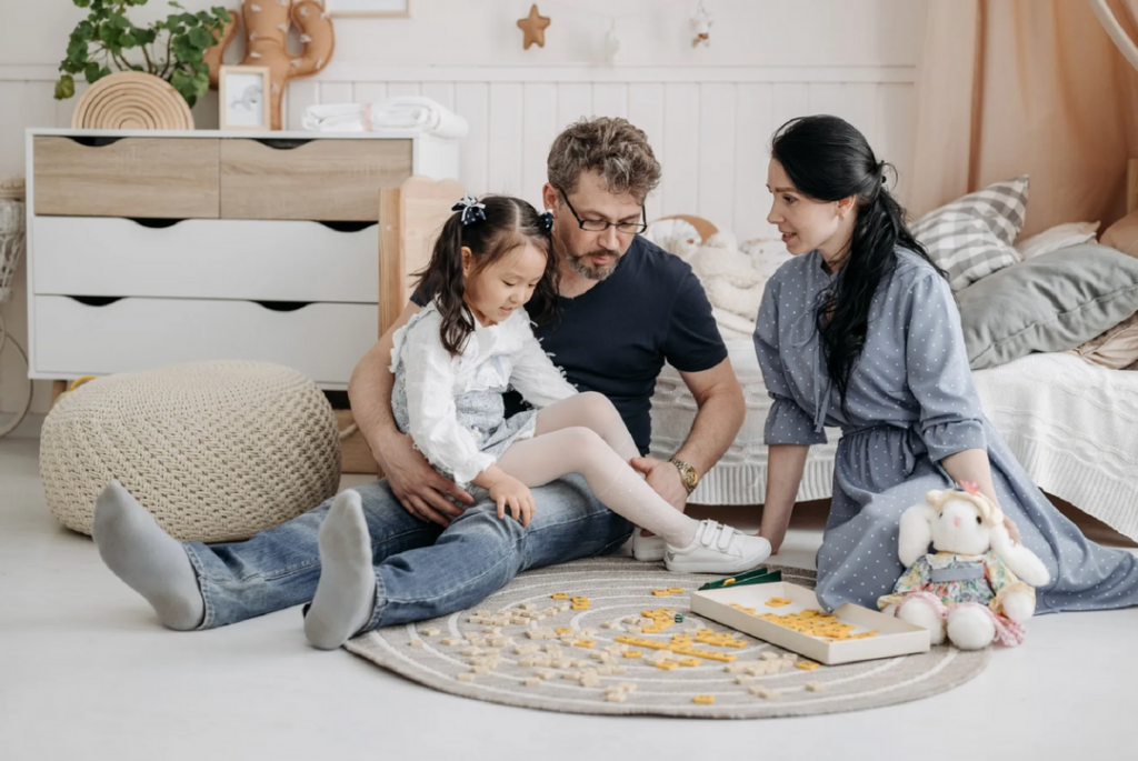 Cozakos and Centeno : Family playing a board game on the floor in a cozy bedroom, with parents helping their young daughter with the game pieces.