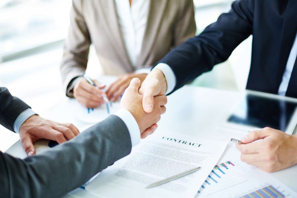 Cozakos and Centeno : Two business professionals shaking hands over a table with a contract and financial charts, signaling agreement or partnership.