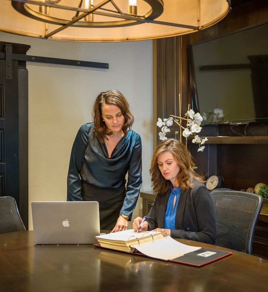 Cozakos and Centeno : Two women collaborating at a desk with a laptop and documents in an office setting.