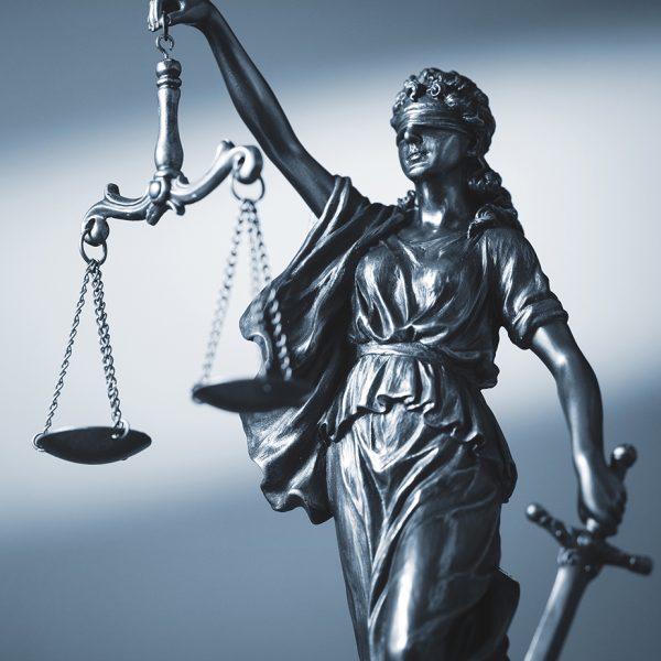 Cozakos and Centeno : Close-up of the statue of lady justice, featuring a blindfolded figure holding balanced scales and a sword, against a blue background.