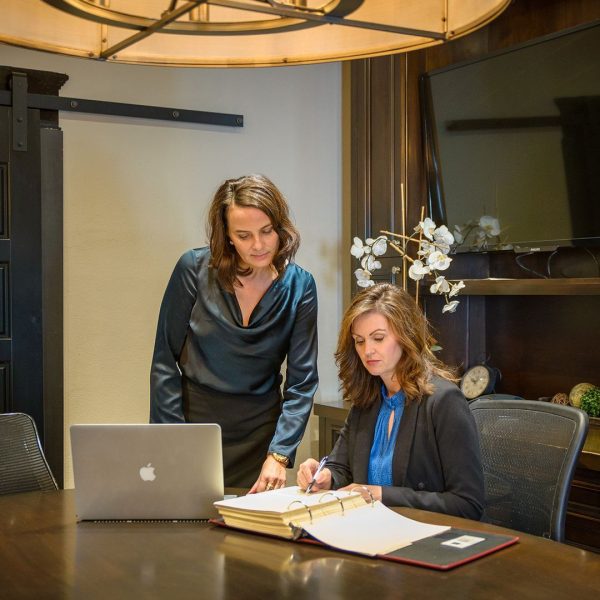 Cozakos and Centeno : Two women collaborating at a desk with a laptop and documents in an office setting.